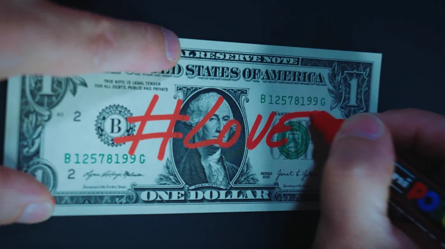 Les mains de Damien Thevenet écrivent « #Love » au feutre rouge sur un billet de un dollar