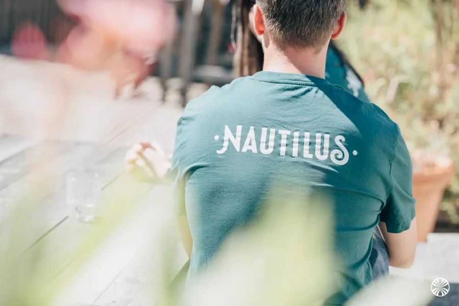 De dos, un membre de l’équipe en t-shirt vert floqué « Nautilus », assis en terrasse au soleil